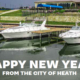 Several white motorboats docked at a marina on calm water, with grassy terraces and buildings in the background. Large white text across the bottom reads, ‘HAPPY NEW YEAR FROM THE CITY OF HEATH.