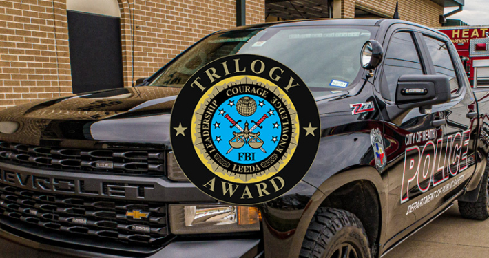 Black police pickup truck parked outside a brick building, marked “City of Heath Police,” with emergency lights and a fire truck visible in the background; an FBI “Trilogy Award” emblem is overlaid prominently in the center of the image.