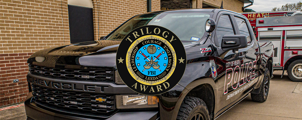 Black police pickup truck parked outside a brick building, marked “City of Heath Police,” with emergency lights and a fire truck visible in the background; an FBI “Trilogy Award” emblem is overlaid prominently in the center of the image.
