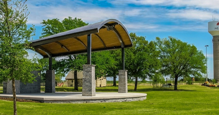 The new outdoor pavilion at Towne Center Park, with a curved metal roof supported by brick columns sits on a concrete slab in a grassy park. Trees surround the area, and the south water tower stands in the background under a clear blue sky.