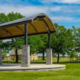 The new outdoor pavilion at Towne Center Park, with a curved metal roof supported by brick columns sits on a concrete slab in a grassy park. Trees surround the area, and the south water tower stands in the background under a clear blue sky.