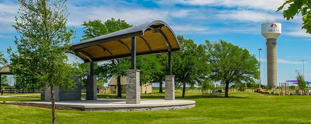 The new outdoor pavilion at Towne Center Park, with a curved metal roof supported by brick columns sits on a concrete slab in a grassy park. Trees surround the area, and the south water tower stands in the background under a clear blue sky.