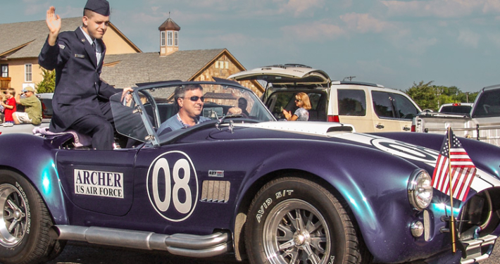 A person in a U.S. Air Force uniform waves from a vintage purple Shelby Cobra convertible during the 2008 Independence Day Parade in Heath, Texas, with American flags on the car and spectators watching from the roadside.