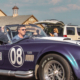 A person in a U.S. Air Force uniform waves from a vintage purple Shelby Cobra convertible during the 2008 Independence Day Parade in Heath, Texas, with American flags on the car and spectators watching from the roadside.