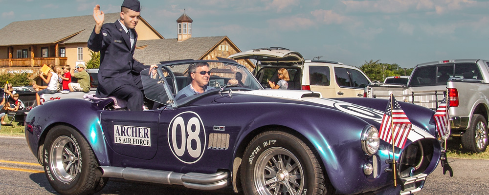A person in a U.S. Air Force uniform waves from a vintage purple Shelby Cobra convertible during the 2008 Independence Day Parade in Heath, Texas, with American flags on the car and spectators watching from the roadside.