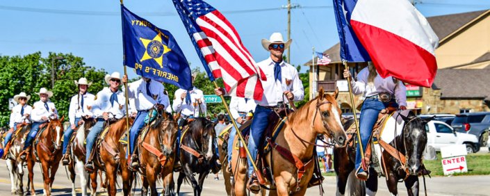 Members of the Rockwall Sheriff's Posse ride horses in the July 4th parade in Heath, Texas. Riders dressed in white shirts, blue neckerchiefs, jeans, and cowboy hats carry the American flag, Texas flag, and posse banner while proceeding down a sunny parade route lined with spectators and vehicles.