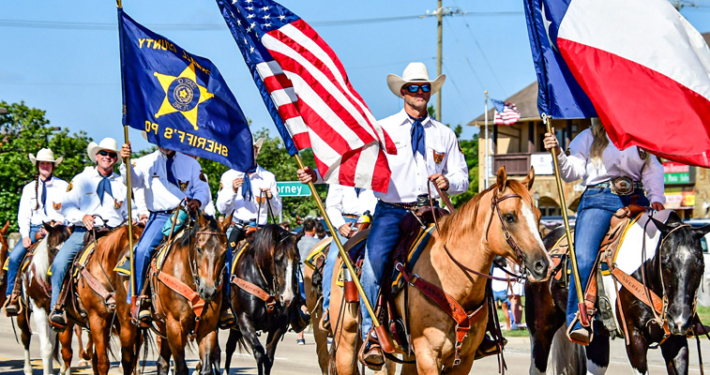 Members of the Rockwall Sheriff's Posse ride horses in the July 4th parade in Heath, Texas. Riders dressed in white shirts, blue neckerchiefs, jeans, and cowboy hats carry the American flag, Texas flag, and posse banner while proceeding down a sunny parade route lined with spectators and vehicles.