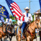 Members of the Rockwall Sheriff's Posse ride horses in the July 4th parade in Heath, Texas. Riders dressed in white shirts, blue neckerchiefs, jeans, and cowboy hats carry the American flag, Texas flag, and posse banner while proceeding down a sunny parade route lined with spectators and vehicles.