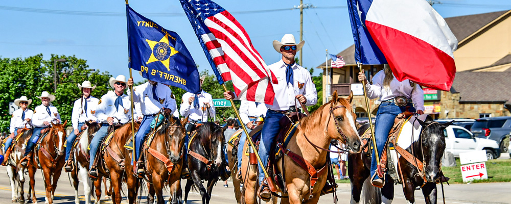 Members of the Rockwall Sheriff's Posse ride horses in the July 4th parade in Heath, Texas. Riders dressed in white shirts, blue neckerchiefs, jeans, and cowboy hats carry the American flag, Texas flag, and posse banner while proceeding down a sunny parade route lined with spectators and vehicles.