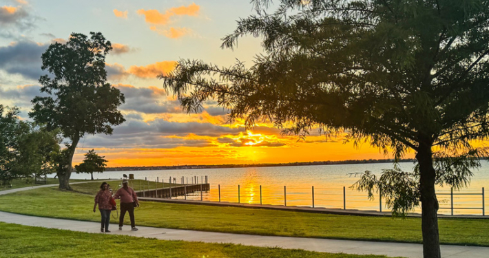 A peaceful sunset at Terry Park in Heath, Texas, with two people walking along a paved lakeside trail, green parkland, trees, and a calm lake reflecting the golden-orange sky.