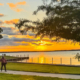 A peaceful sunset at Terry Park in Heath, Texas, with two people walking along a paved lakeside trail, green parkland, trees, and a calm lake reflecting the golden-orange sky.
