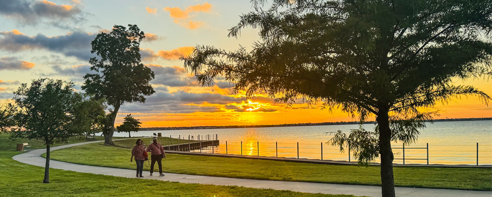 A peaceful sunset at Terry Park in Heath, Texas, with two people walking along a paved lakeside trail, green parkland, trees, and a calm lake reflecting the golden-orange sky.
