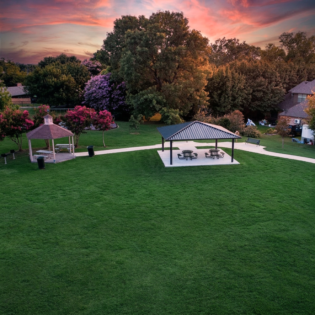 An elevated view of Antigua Bay park at sunset. A paved walking path winds past a small hexagonal gazebo on the left and a larger, open-air pavilion with a dark, gabled roof and picnic tables on the right.