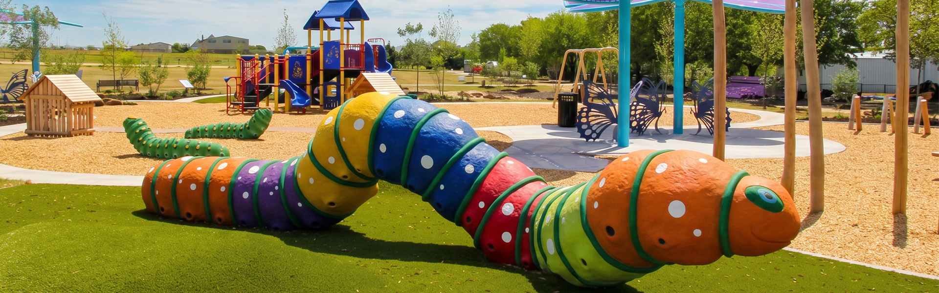 Colorful caterpillar-shaped playground tunnel sculpture on artificial turf at Towne Center Park in Heath, Texas, with playground equipment, walking paths, and shaded seating areas in the background.