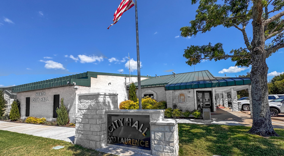 Exterior view of Heath City Hall at 200 Laurence Drive in Heath, Texas, featuring the city hall building, entrance sign, American flag, large oak tree, and landscaped grounds under a clear blue sky.