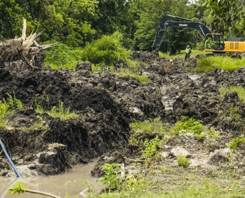 A daytime outdoor scene of heavy construction in a wooded area. A deep, heavily churned trench filled with dark mud and murky water runs through the center, with a blue pipe partially visible in the water in the foreground. On the left bank of the mud sits a large uprooted tree stump with exposed roots. In the background, a yellow John Deere excavator is actively working in the trench, while a construction worker wearing a hard hat and high-visibility yellow-green clothing stands nearby. Dense green trees surround the site.