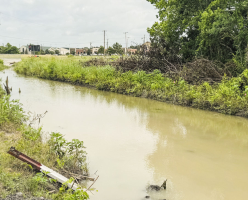 A wide drainage ditch filled with murky, brown water flowing between grassy banks. The left bank is covered in green weeds and vegetation, with a broken red and white post lying in the mud near the water's edge. The right bank features dense green trees and a large pile of cleared, brown brush. In the background, a concrete bridge structure spans the waterway, leading to a street lined with commercial buildings and utility poles under a cloudy, overcast sky.