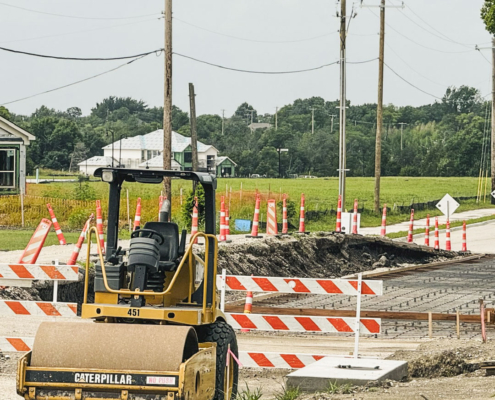 A daytime view of a road construction site. In the foreground, a yellow Caterpillar road roller with the number "451" sits parked on rough dirt. Just behind the roller, orange and white striped barricades block off a section of the road where the surface has been removed and a metal rebar grid is laid out, prepped for a concrete pour. A long line of orange construction cones stretches along the side of the road and into the adjacent green field. In the background, wooden utility poles carry power lines across the scene, and houses under construction are visible amongst dense green trees under an overcast sky.