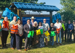 A group of about 20 community members, local officials, first responders, and law enforcement personnel stand together at a waterfront park in front of a newly installed life jacket loaner station. Several people hold brightly colored life jackets, while additional life jackets hang on the station behind them. Trees and a lake are visible in the background on a sunny day, highlighting a community water safety initiative.