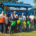 A group of about 20 community members, local officials, first responders, and law enforcement personnel stand together at a waterfront park in front of a newly installed life jacket loaner station. Several people hold brightly colored life jackets, while additional life jackets hang on the station behind them. Trees and a lake are visible in the background on a sunny day, highlighting a community water safety initiative.