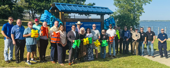 LifeJacketStationTerry1 A group of about 20 community members, local officials, first responders, and law enforcement personnel stand together at a waterfront park in front of a newly installed life jacket loaner station. Several people hold brightly colored life jackets, while additional life jackets hang on the station behind them. Trees and a lake are visible in the background on a sunny day, highlighting a community water safety initiative.