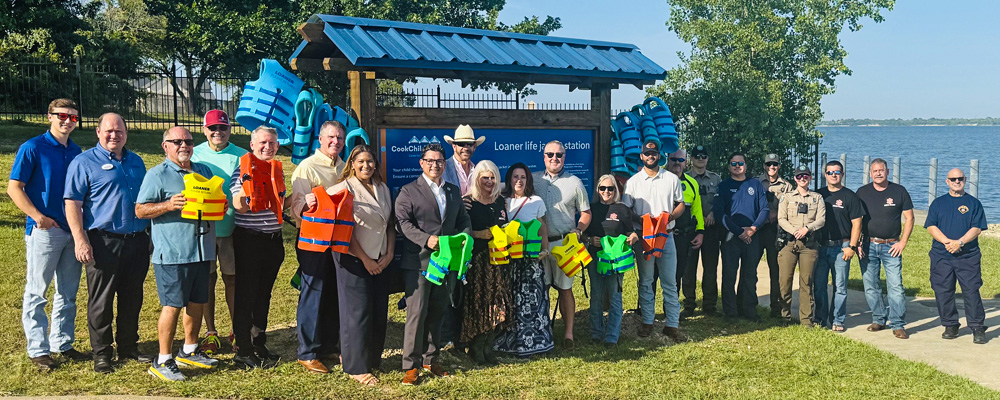 A group of about 20 community members, local officials, first responders, and law enforcement personnel stand together at a waterfront park in front of a newly installed life jacket loaner station. Several people hold brightly colored life jackets, while additional life jackets hang on the station behind them. Trees and a lake are visible in the background on a sunny day, highlighting a community water safety initiative.