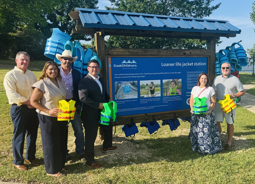 Six people stand around a newly installed life jacket loaner station at a lakeside park, each holding brightly colored child or adult life jackets. The station is stocked with blue life jackets and features a large instructional sign explaining how to choose and properly fit a U.S. Coast Guard-approved life jacket. Trees and the lake are visible in the background on a sunny day, emphasizing the community's water safety initiative.