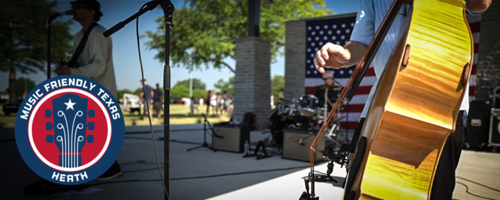 MusicFriendlyTx Musicians perform on stage at the amphitheater in Towne Center Park in Heath, Texas, during an outdoor community celebration. A close-up of an upright bass is shown in the foreground, with a drummer, microphone stands, and an American flag visible on stage. A "Music Friendly Texas – Heath" logo appears in the lower-left corner.