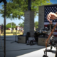 Musicians perform on stage at the amphitheater in Towne Center Park in Heath, Texas, during an outdoor community celebration. A close-up of an upright bass is shown in the foreground, with a drummer, microphone stands, and an American flag visible on stage. A "Music Friendly Texas – Heath" logo appears in the lower-left corner.