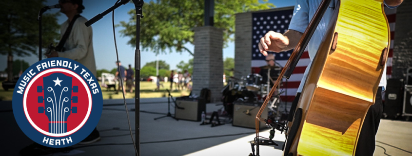Musicians perform on stage at the amphitheater in Towne Center Park in Heath, Texas, during an outdoor community celebration. A close-up of an upright bass is shown in the foreground, with a drummer, microphone stands, and an American flag visible on stage. A "Music Friendly Texas – Heath" logo appears in the lower-left corner.