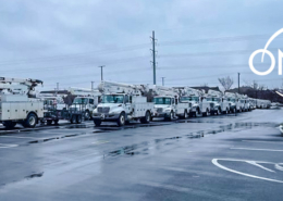 A long row of white Oncor utility bucket trucks parked in a wet lot on an overcast winter day, with patches of snow along the edges and the Oncor logo displayed in the upper right.
