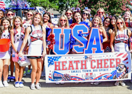 A large group of youth cheerleaders wearing red, white, and blue uniforms pose together during an Independence Day parade. They hold patriotic pom-poms, American flags, and a large "USA" sign behind a banner reading "Heath Cheer – Small Town. Big Spirit." Many participants wear red sunglasses and festive patriotic accessories, celebrating the Fourth of July in a bright outdoor setting.