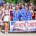 A large group of youth cheerleaders wearing red, white, and blue uniforms pose together during an Independence Day parade. They hold patriotic pom-poms, American flags, and a large "USA" sign behind a banner reading "Heath Cheer – Small Town. Big Spirit." Many participants wear red sunglasses and festive patriotic accessories, celebrating the Fourth of July in a bright outdoor setting.