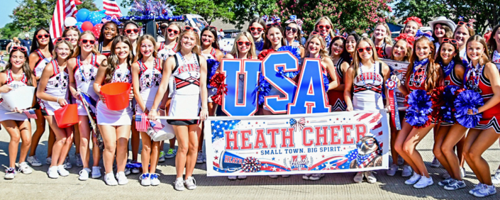 ParadeDerbyWinners A large group of youth cheerleaders wearing red, white, and blue uniforms pose together during an Independence Day parade. They hold patriotic pom-poms, American flags, and a large "USA" sign behind a banner reading "Heath Cheer – Small Town. Big Spirit." Many participants wear red sunglasses and festive patriotic accessories, celebrating the Fourth of July in a bright outdoor setting.