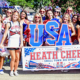 A large group of youth cheerleaders wearing red, white, and blue uniforms pose together during an Independence Day parade. They hold patriotic pom-poms, American flags, and a large "USA" sign behind a banner reading "Heath Cheer – Small Town. Big Spirit." Many participants wear red sunglasses and festive patriotic accessories, celebrating the Fourth of July in a bright outdoor setting.