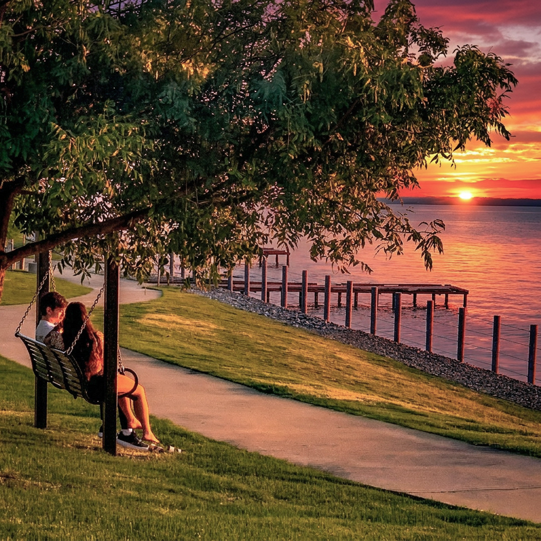 A couple sits together on a hanging swing bench beneath a large, leafy tree next to a paved walking path in Terry Park, Heath. They are looking out over Lake Ray Hubbard toward a vibrant sunset with striking red, orange, and purple clouds. A wooden pier extends into the water in the background, and the golden hour light illuminates the grassy bank.