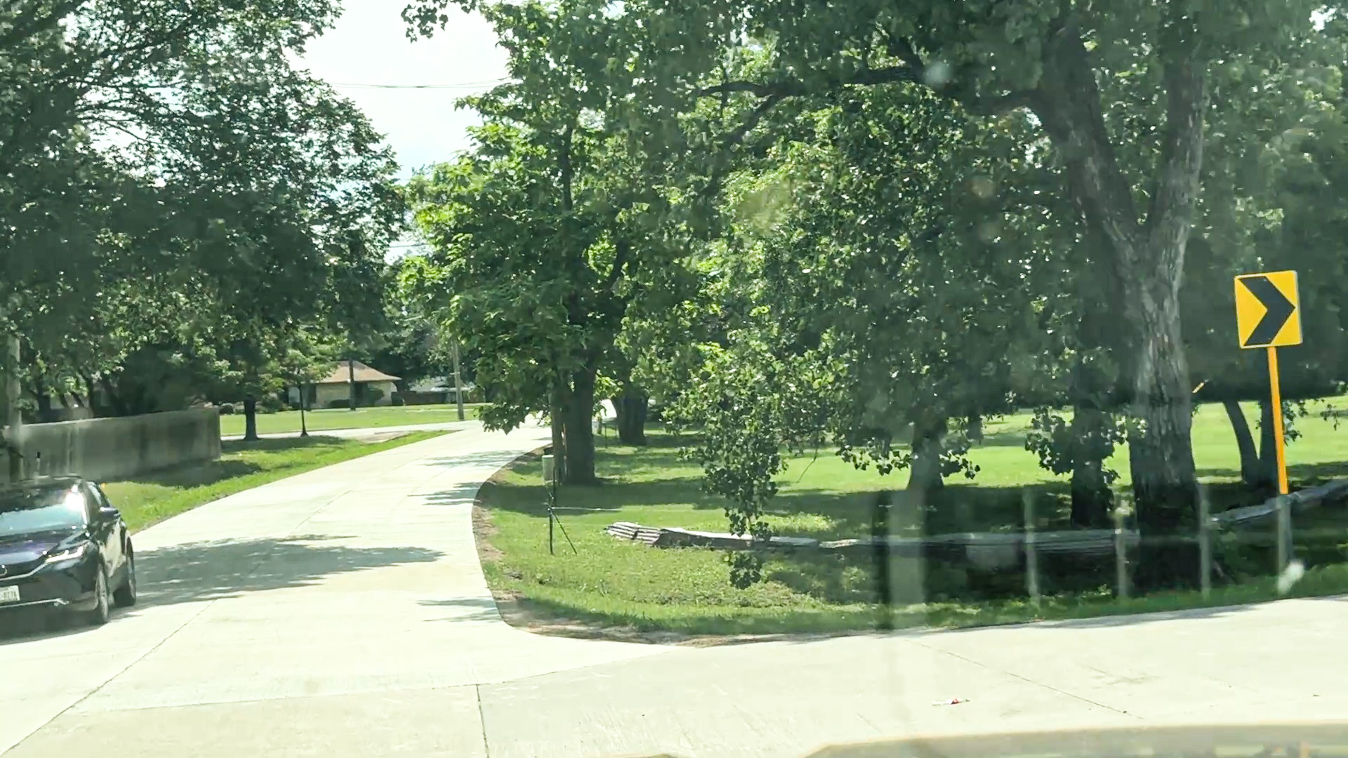 A daytime view from inside a vehicle looking through the windshield at Terry Lane. On the right side, lush green grass and mature trees line the street, accompanied by a yellow chevron road sign pointing right to indicate the curve. On the left side, a dark-colored car is driving toward the viewer alongside a low concrete wall. Houses are visible in the distant background nestled among the trees.