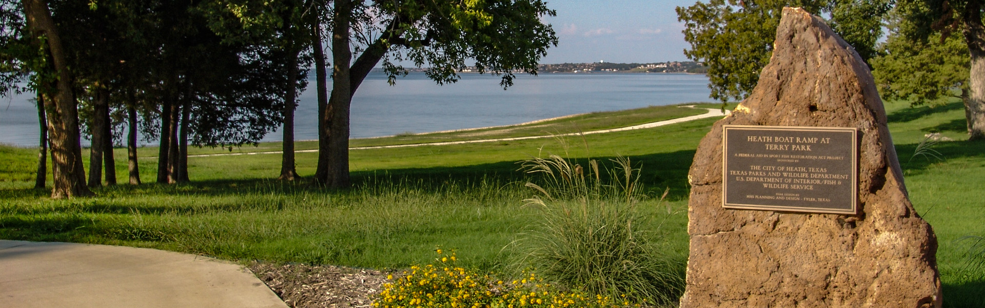 A scenic lakeside view at Terry Park in Heath, Texas, featuring a grassy shoreline, calm water, mature trees casting shade, and a stone monument with a commemorative plaque near a walking path.