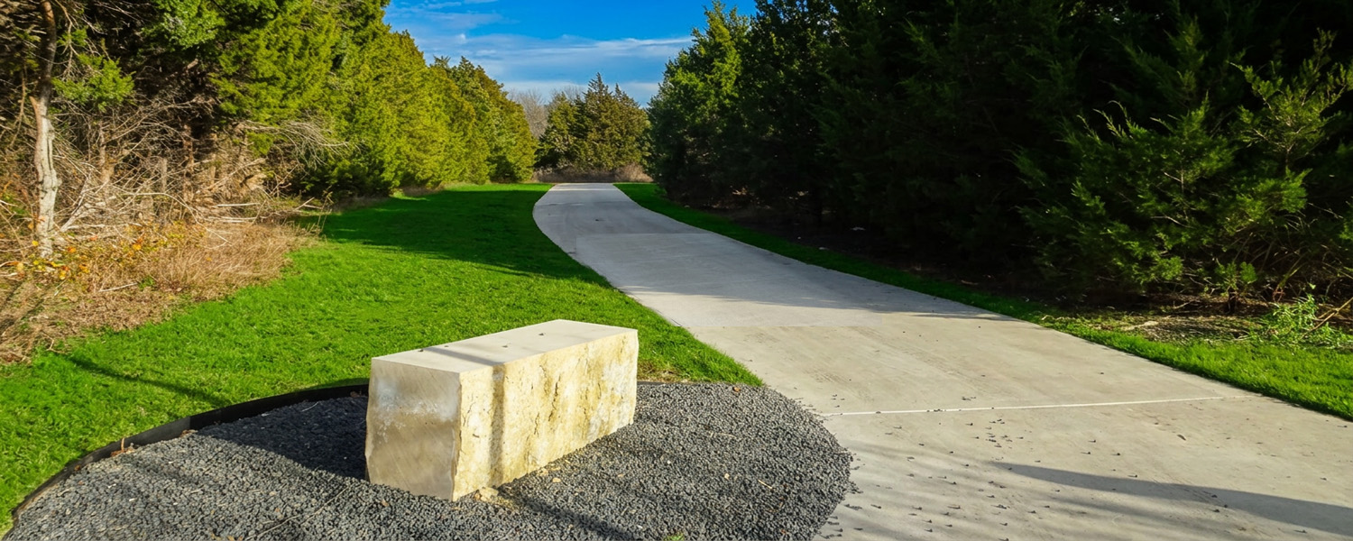 A wide concrete hike and bike trail curves into the distance, bordered by bright green grass and dense, leafy trees under a partly cloudy blue sky. In the foreground beside the path, a large rectangular stone block sits on a patch of dark gravel, serving as a resting bench.